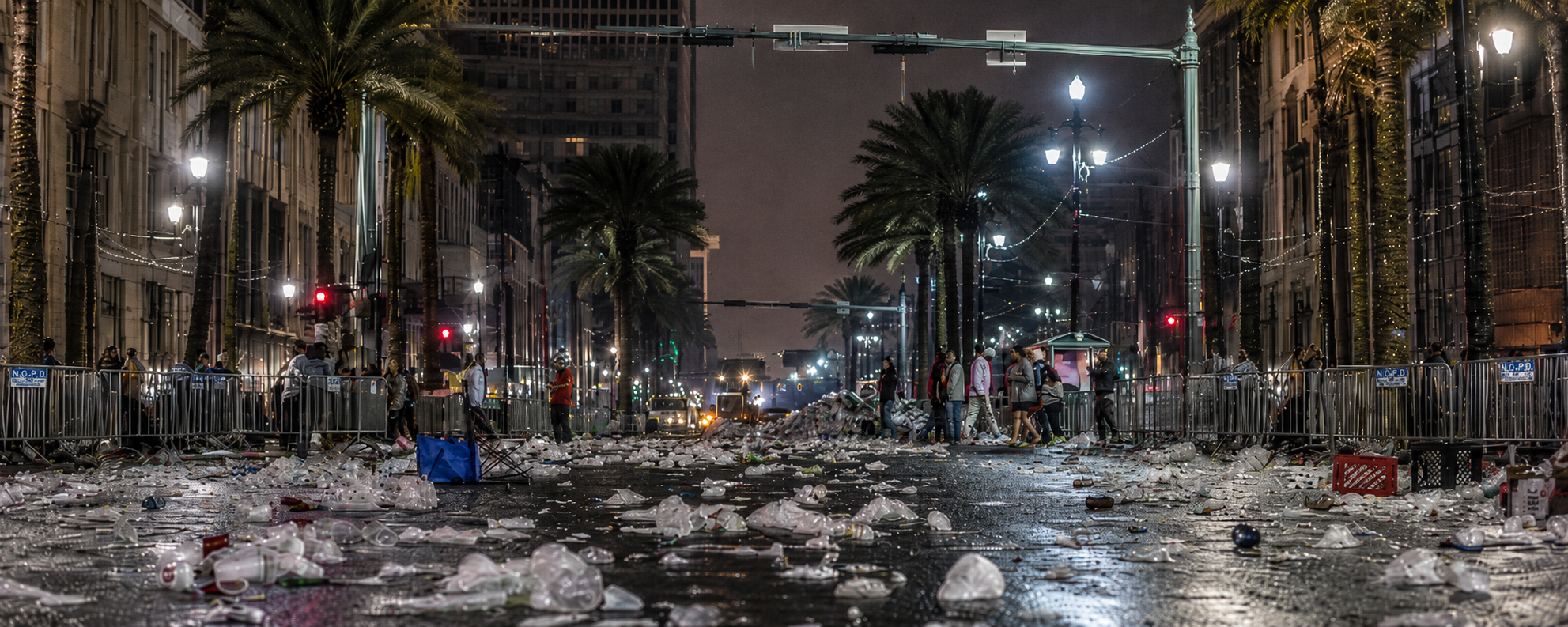 Nighttime city street covered in discarded cups and litter after a public event, with palm trees, police barriers, and wet pavement reflecting streetlights.